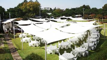 A garden setting with round tables and chairs under white canopies, surrounded by greenery and trees.