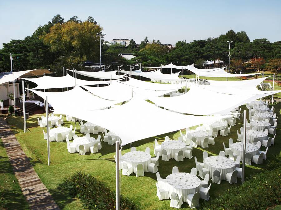 A garden setting with round tables and chairs under white canopies, surrounded by greenery and trees.