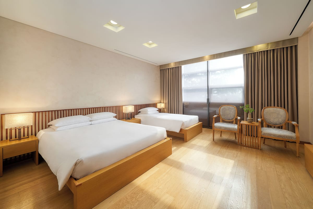 A modern hotel room with two beds, wooden flooring, a seating area with chairs, and a large window with curtains.