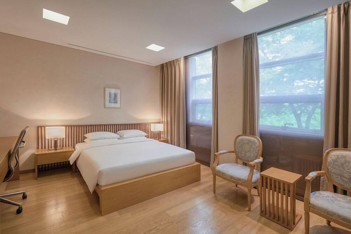 A hotel room with a bed, bedside tables, lamps, a chair, a small table, and a large window with curtains.