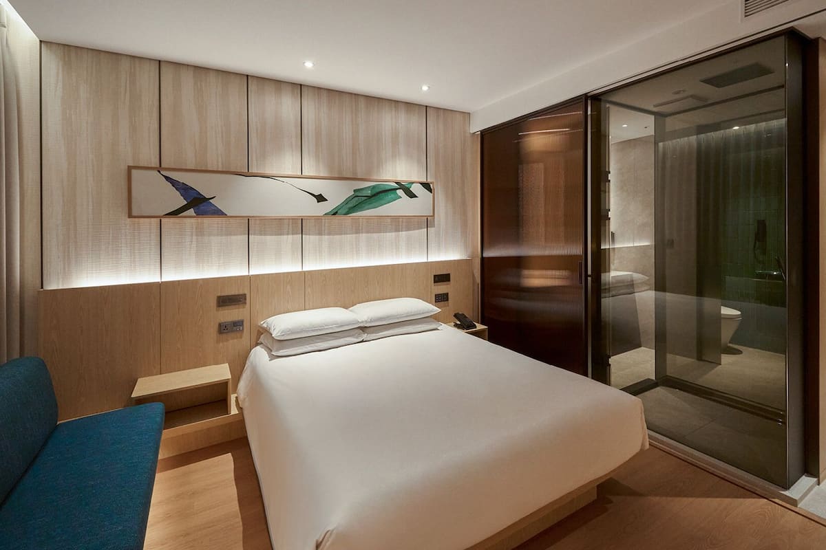 A modern hotel room with a large bed, a desk, a chair, a glass shower enclosure, and a wooden headboard with artwork.