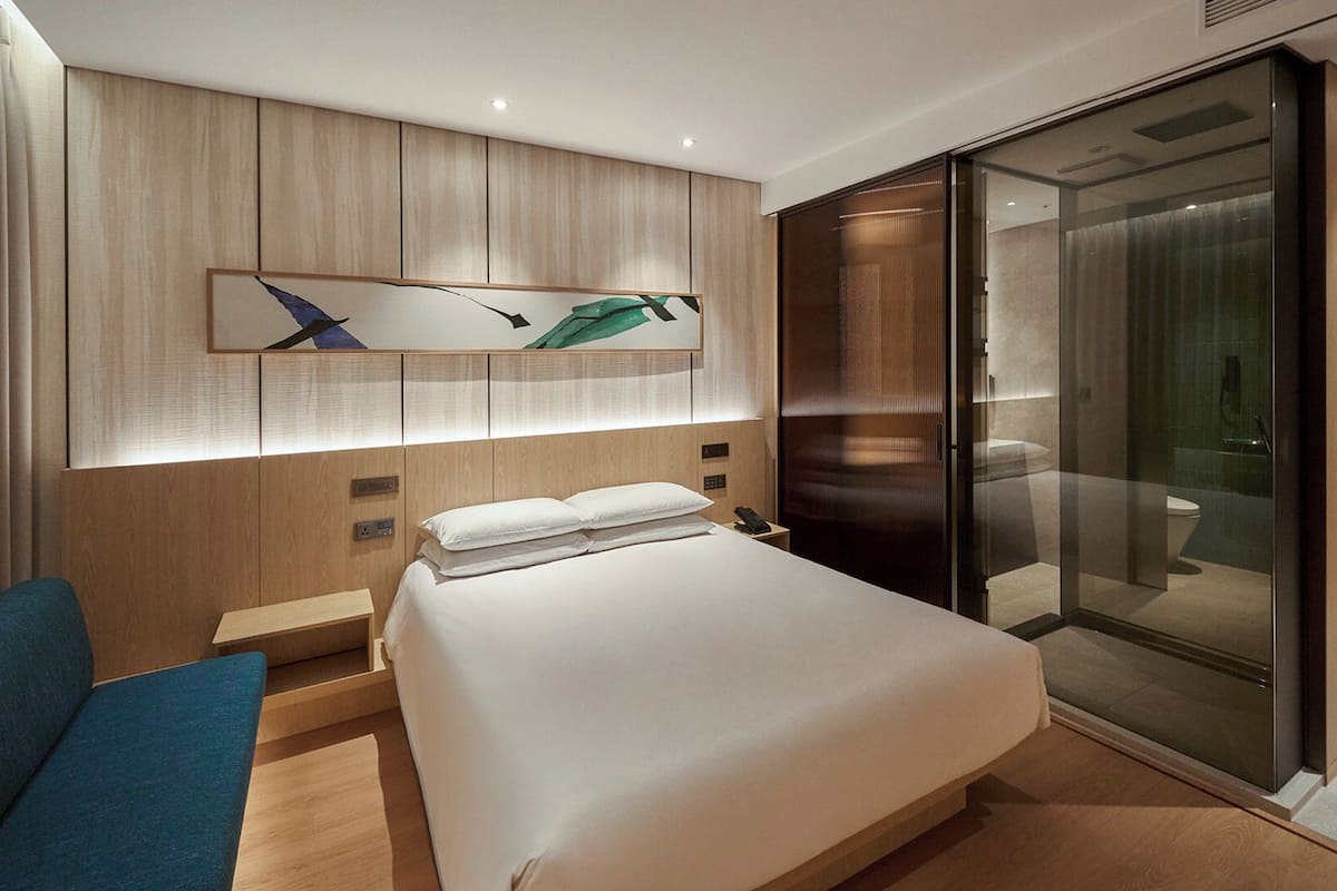 A modern hotel room with a large bed, a desk, a chair, a glass shower enclosure, and a wooden headboard with artwork.