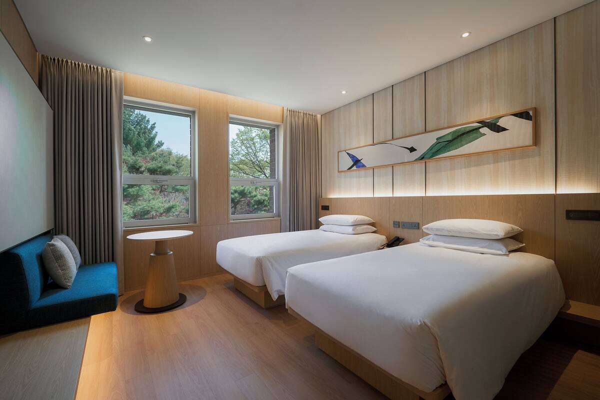 A modern hotel room with two beds, a small round table, a sofa, and a large window with curtains.