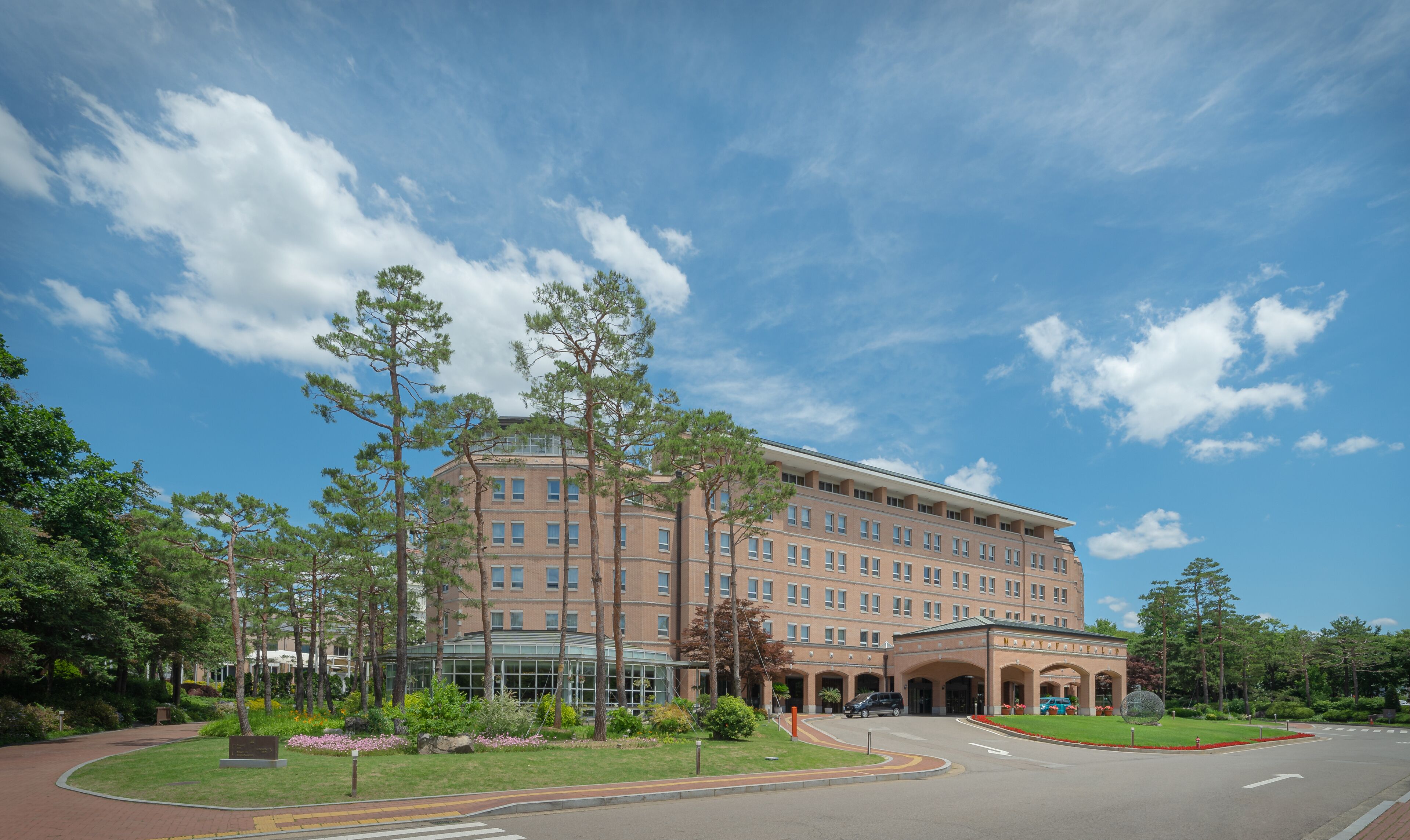 A large brick building with multiple floors, a central entrance with arches, and a landscaped area with trees and shrubs.