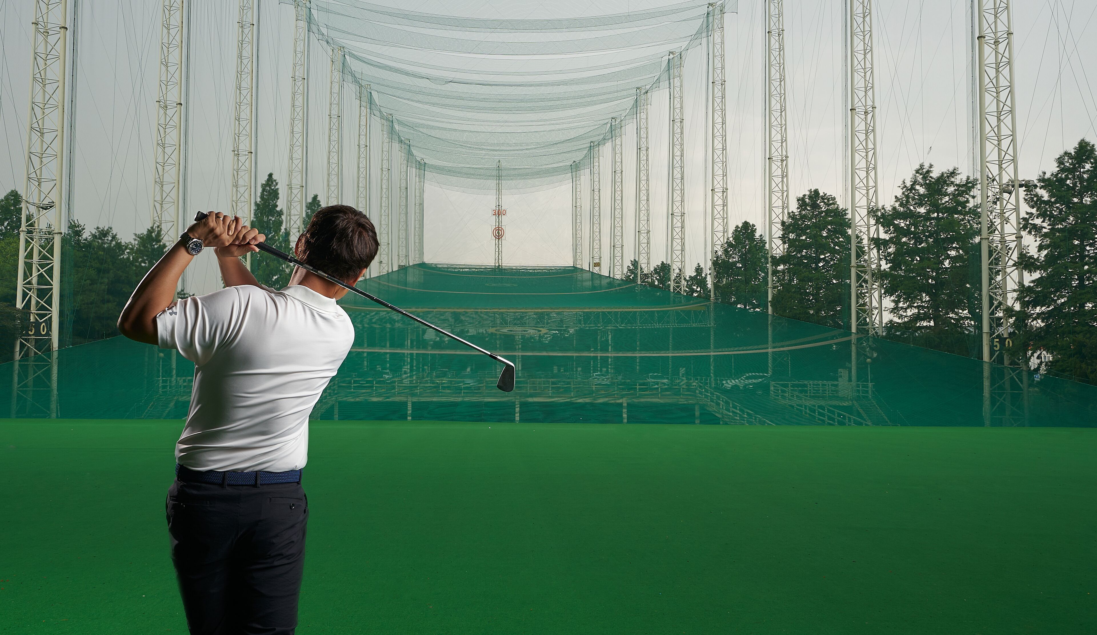 A golfer in a white shirt and dark pants on a green golf course with a netted backdrop and tall poles.