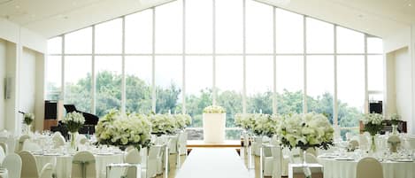 A large hall with a long white table and chairs, large windows, and a piano.