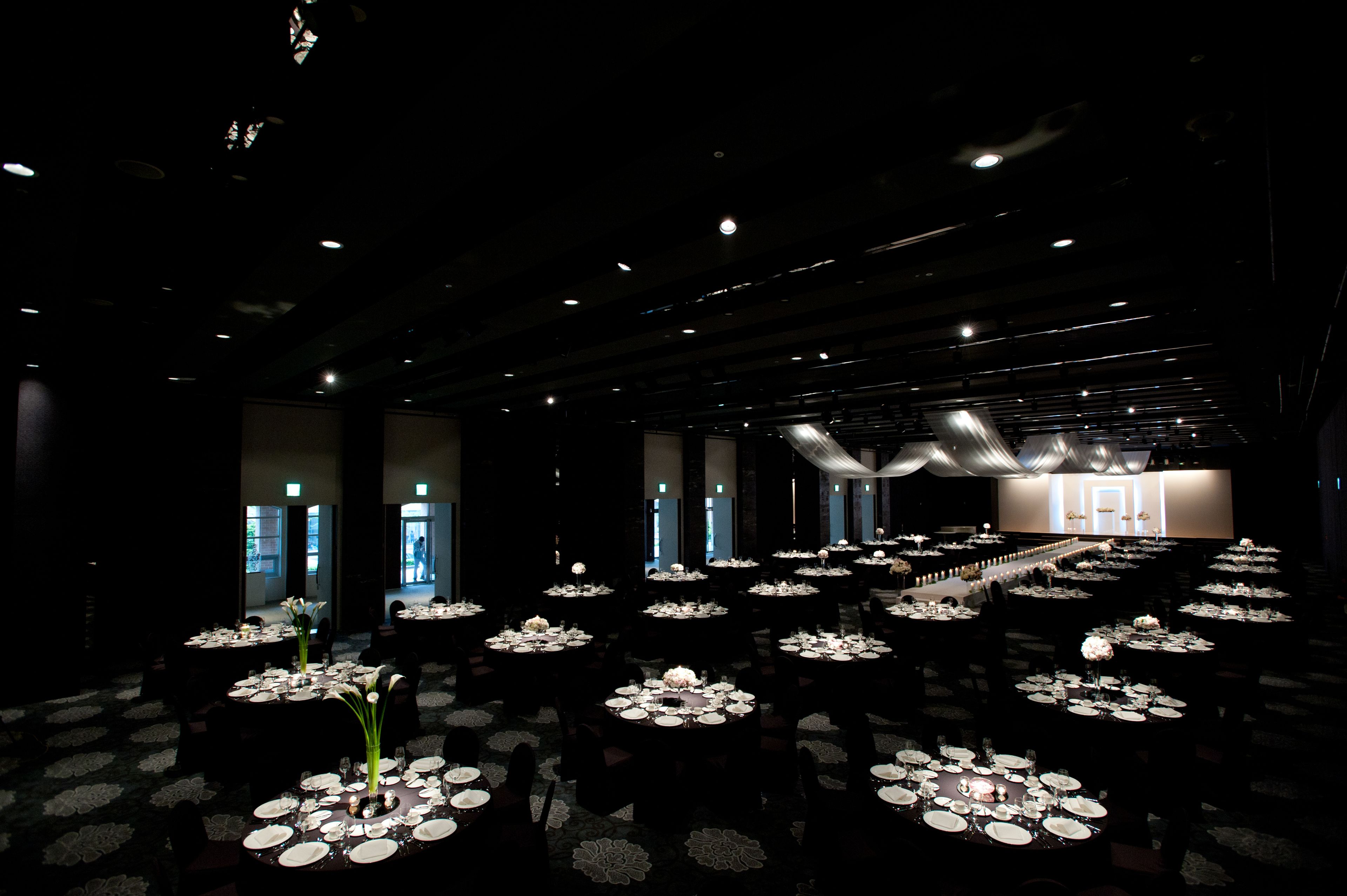 A large, dimly lit banquet hall with multiple round tables set for dining, featuring white tablecloths and floral centerpieces.