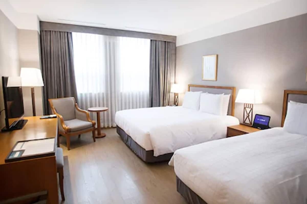 A hotel room with two beds, a desk, a chair, a small table, and a laptop on a nightstand.