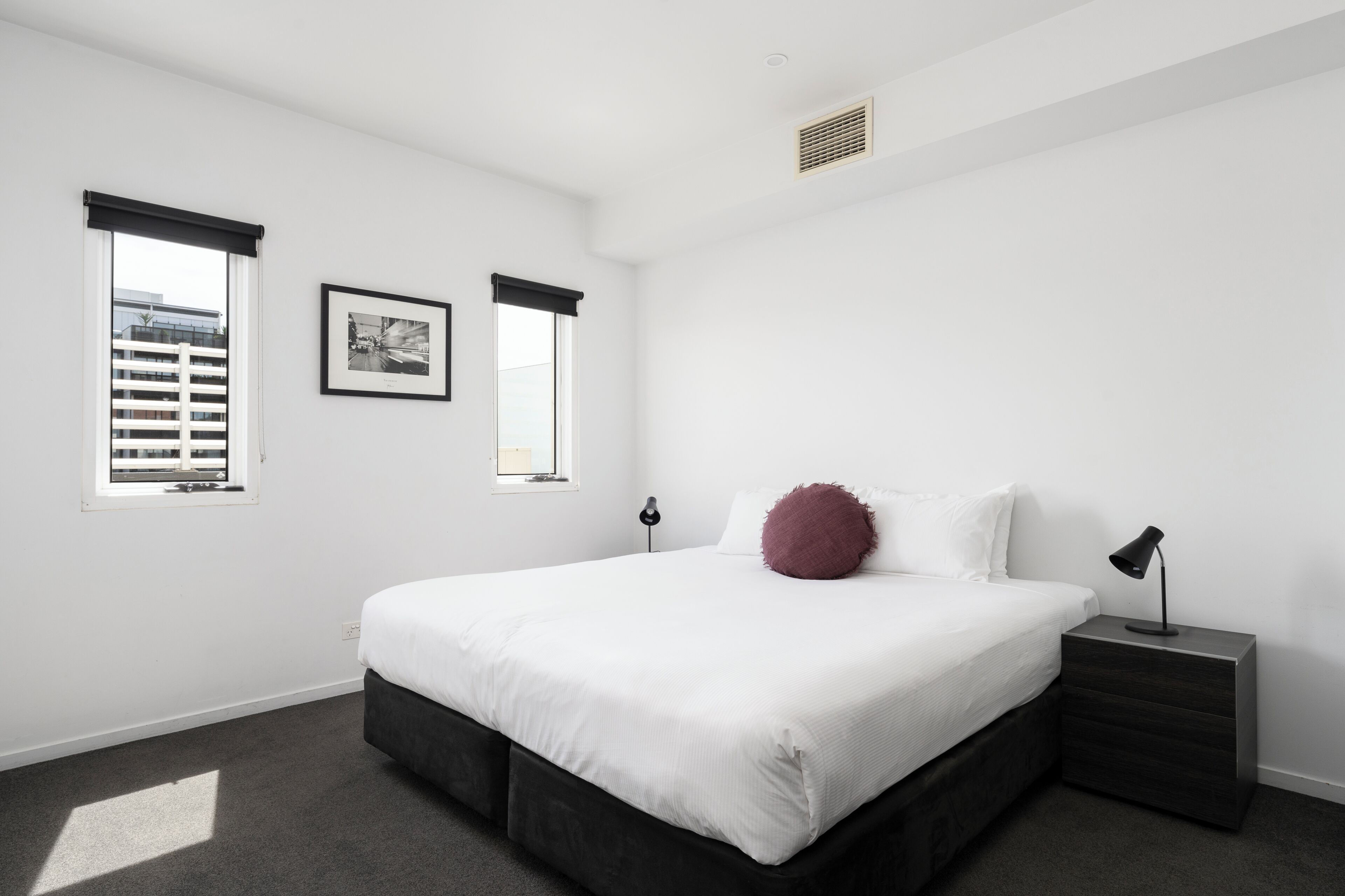 A bedroom with a bed, bedside table, lamp, window, and picture on the wall.