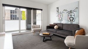 Una sala de estar moderna con un sofá gris, una mesa de centro redonda y una silla. Hay un ventanal con vistas a un edificio y zonas verdes.