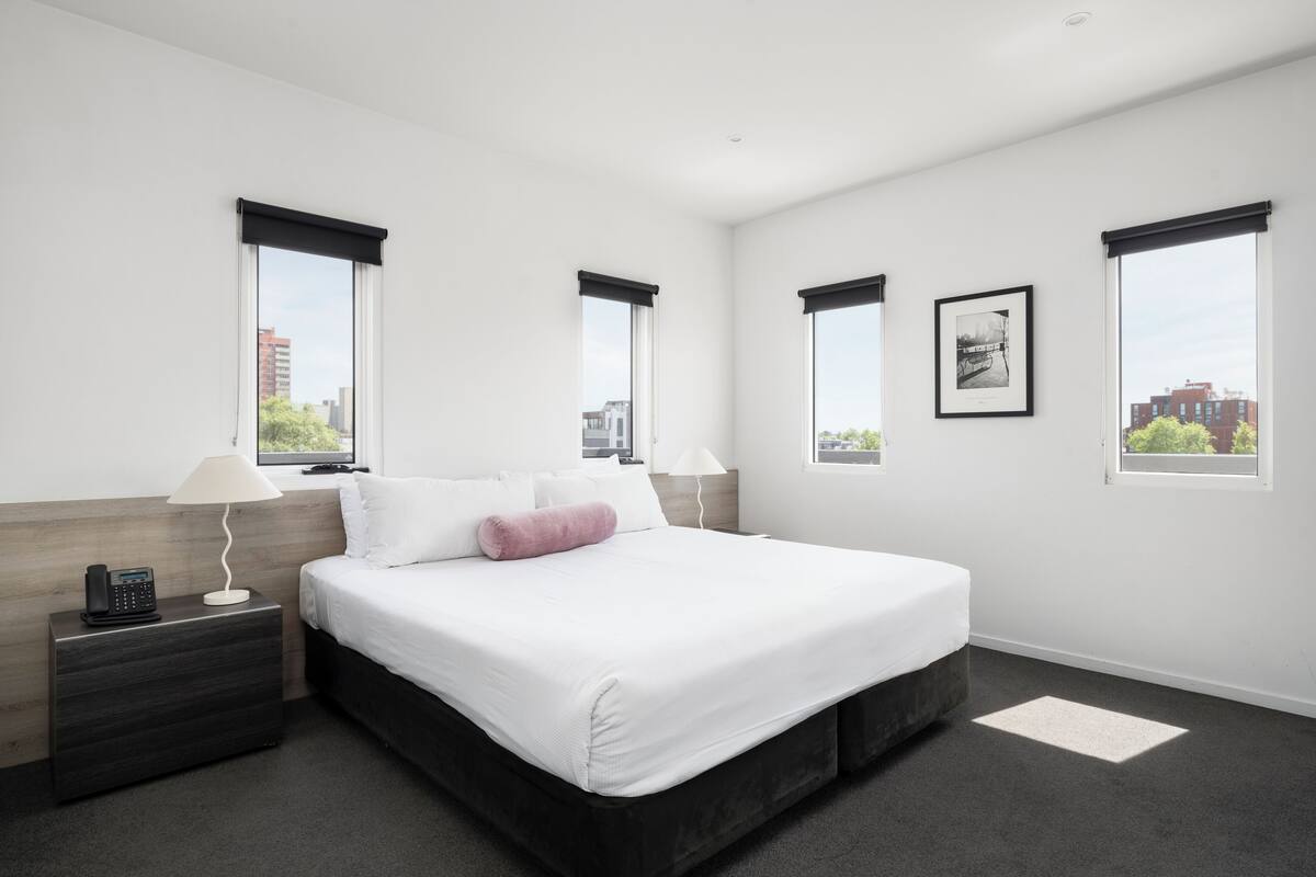 A modern hotel room with a large bed, a bedside table with a lamp, and a city view through the windows.