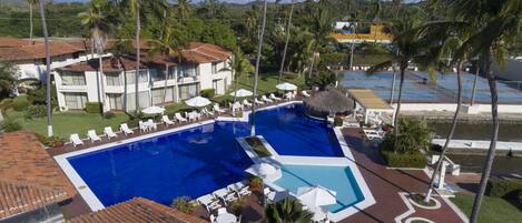 Un resort con una amplia piscina rodeada de sillones de descanso y sombrillas, palmeras y un edificio con techo de tejas.
