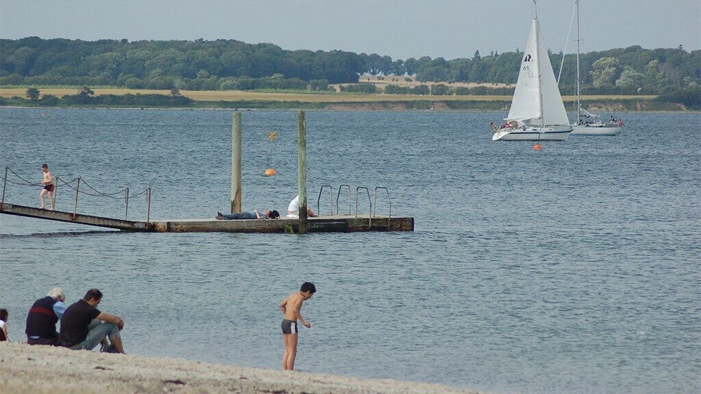 A lakeside scene with a dock extending into the water, a sailboat, and people engaging in various activities.