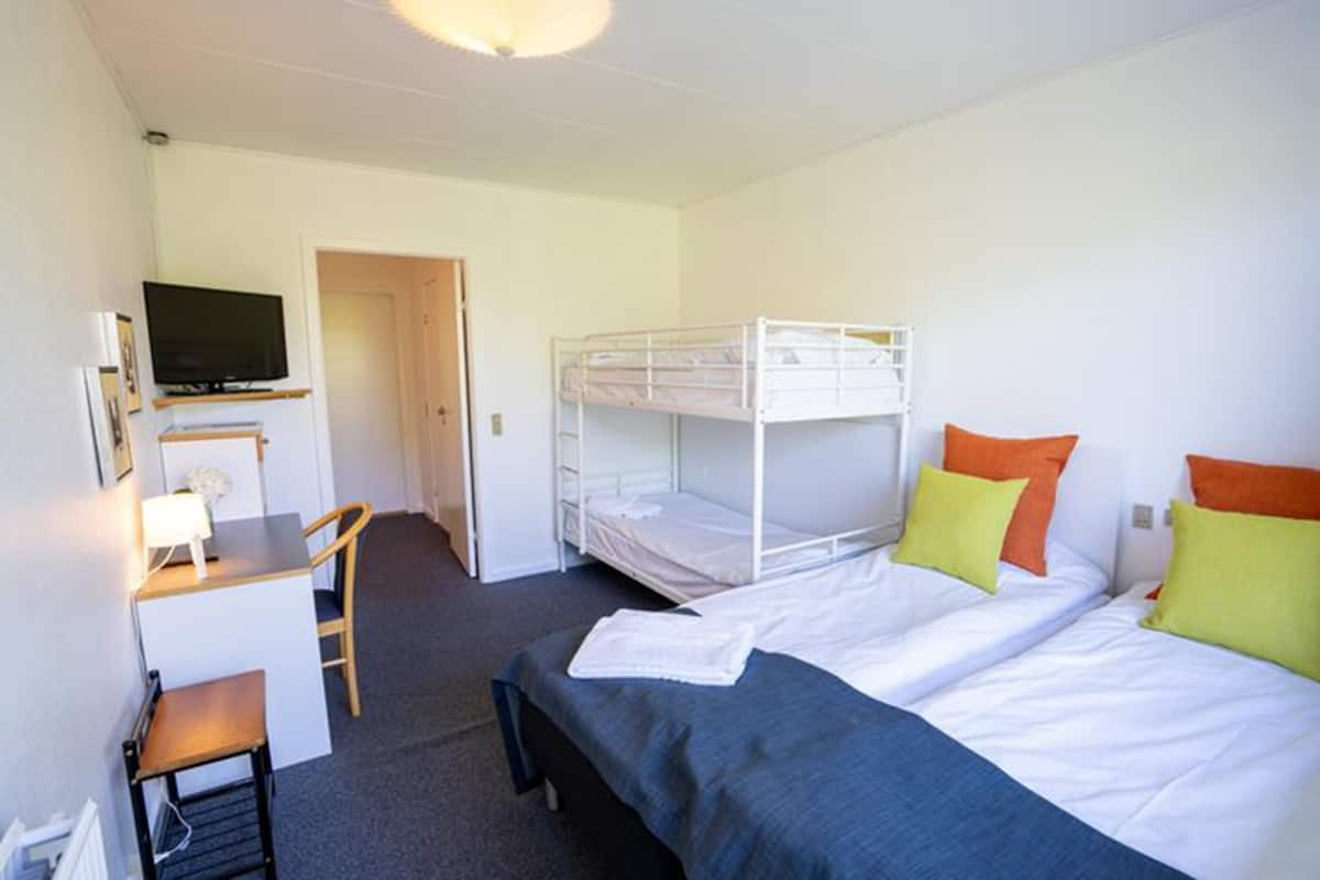 A room with bunk beds, a desk with a chair, a television, and a small table with a lamp.