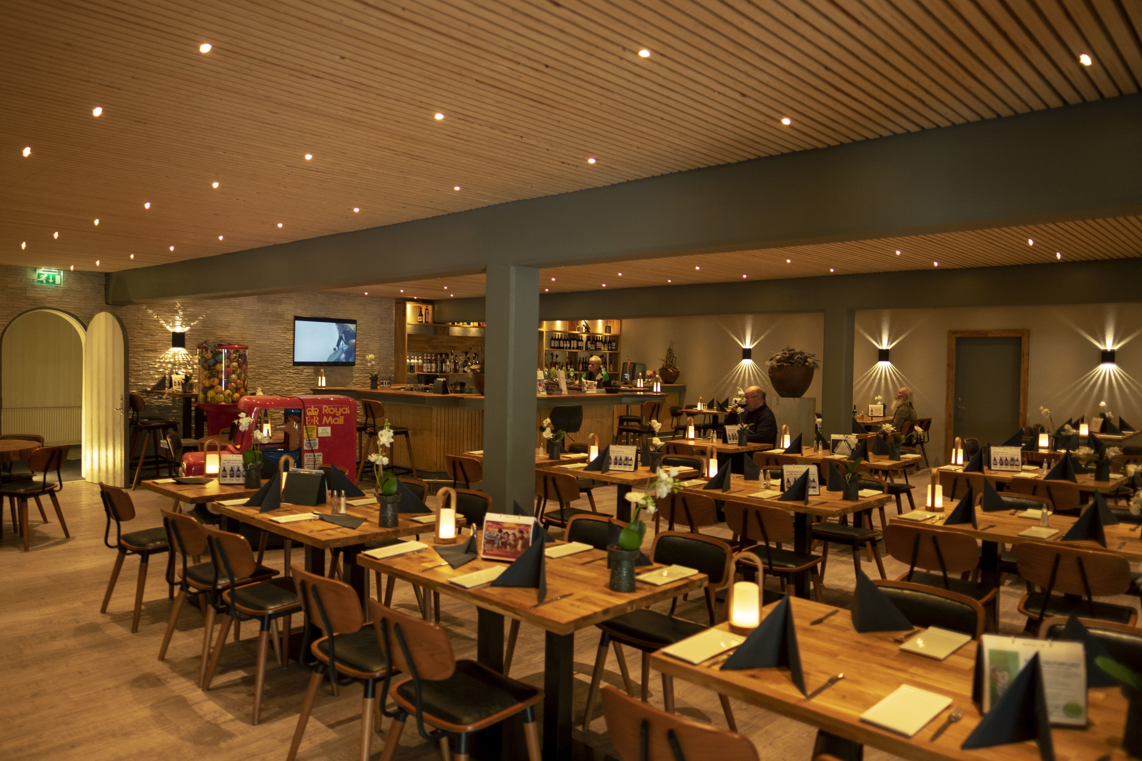 Un restaurante con mesas y sillas de madera, una zona de bar con televisor y diversas plantas y adornos.