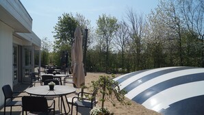 An outdoor patio with round tables and chairs, a large umbrella, and a distinctive striped structure. - Hotel Pejsegården (Braedstrup)