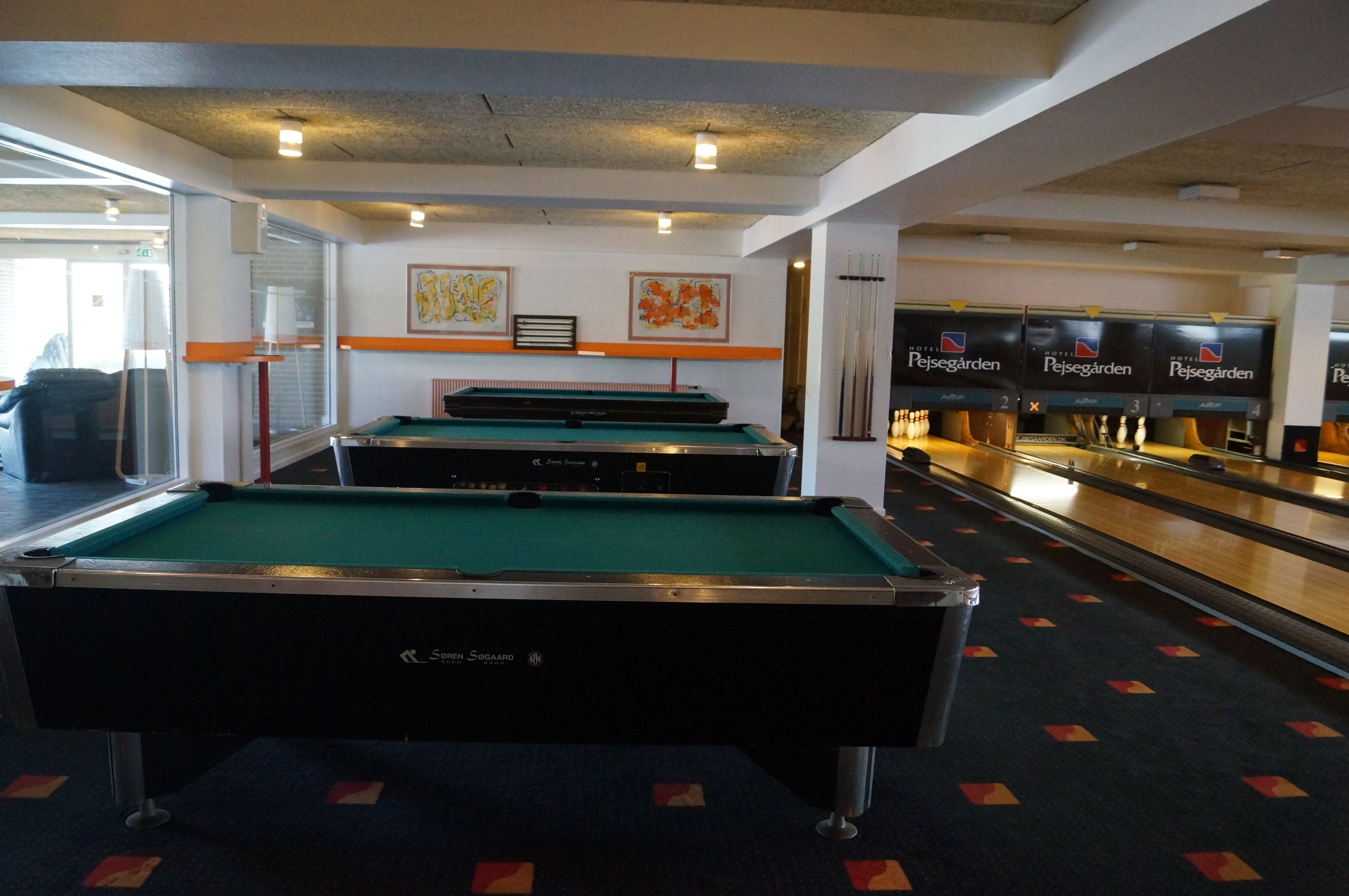A pool table with a green felt surface, surrounded by a carpeted floor with red and yellow diamond patterns. There are two framed artworks on the wall above the table.