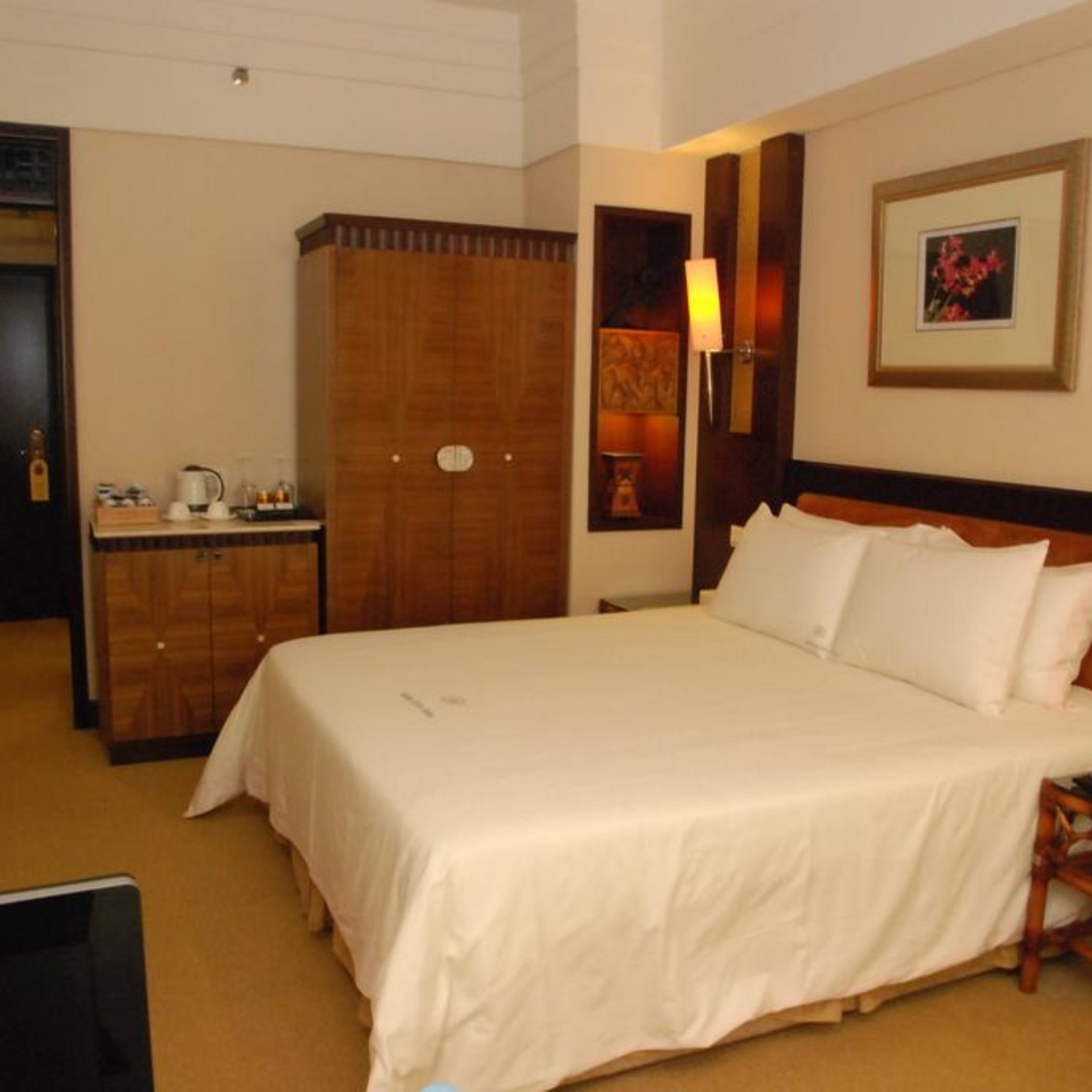 A hotel room with a large bed, a wooden wardrobe, a nightstand with a lamp, and a framed picture on the wall.