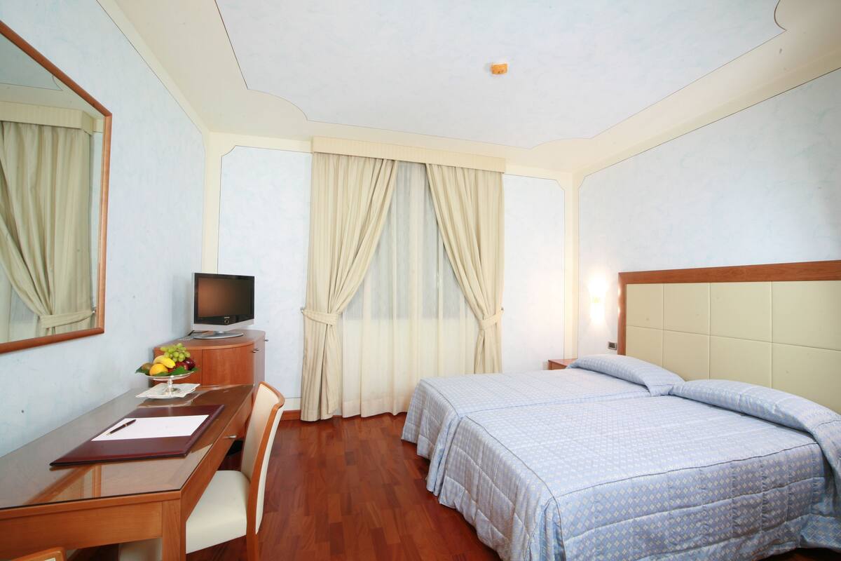 Habitación de hotel con escritorio de madera, televisor, cama con colcha azul y ventana con cortinas.