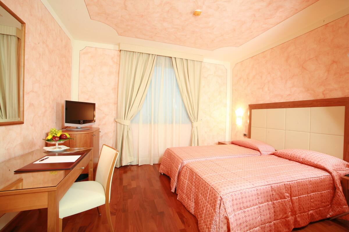 Habitación de hotel con escritorio de madera, televisor, cama con ropa de cama rosa y ventana con cortinas.