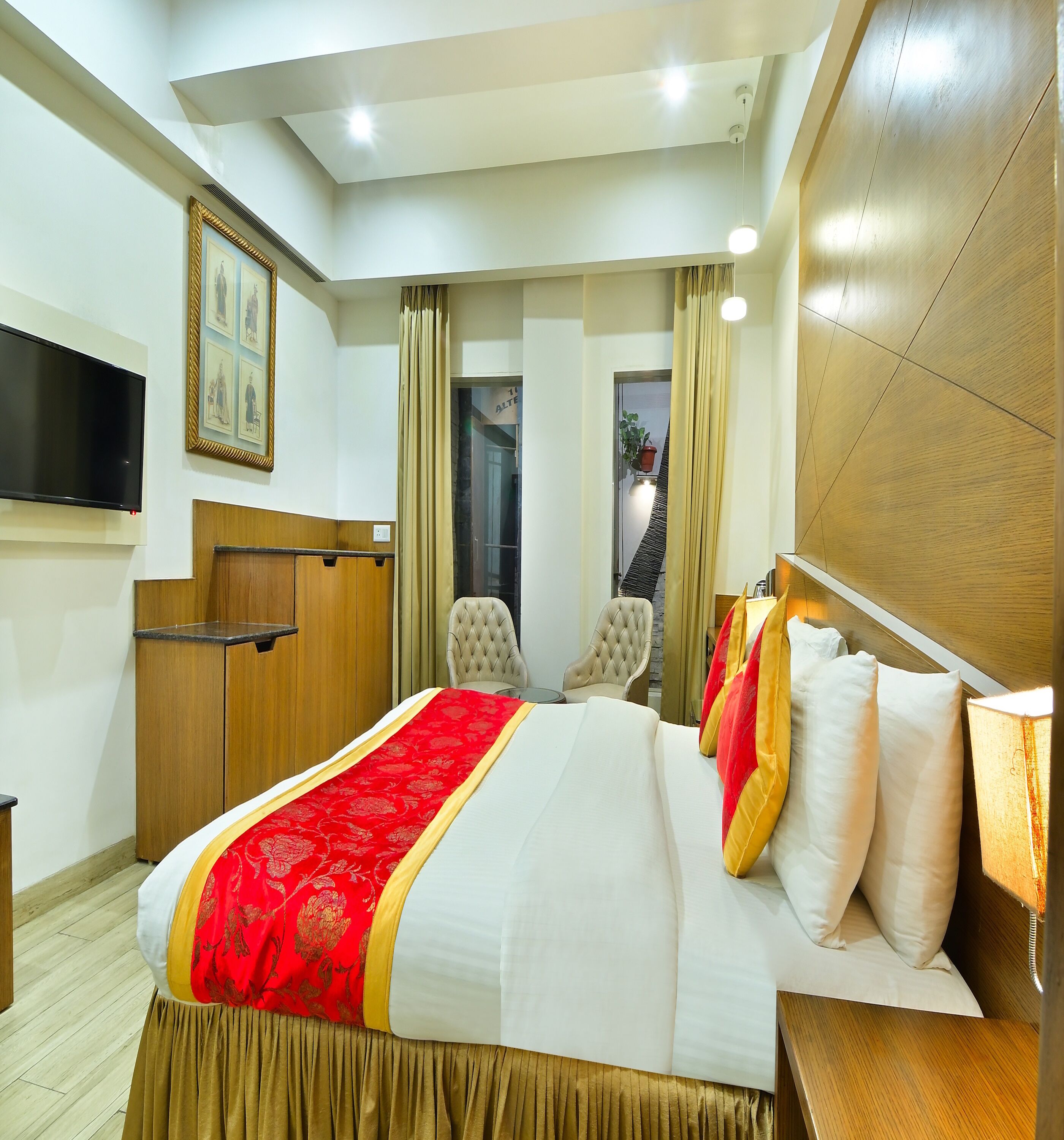 A hotel room with a large bed, a TV mounted on the wall, a wooden headboard, and a window with curtains.