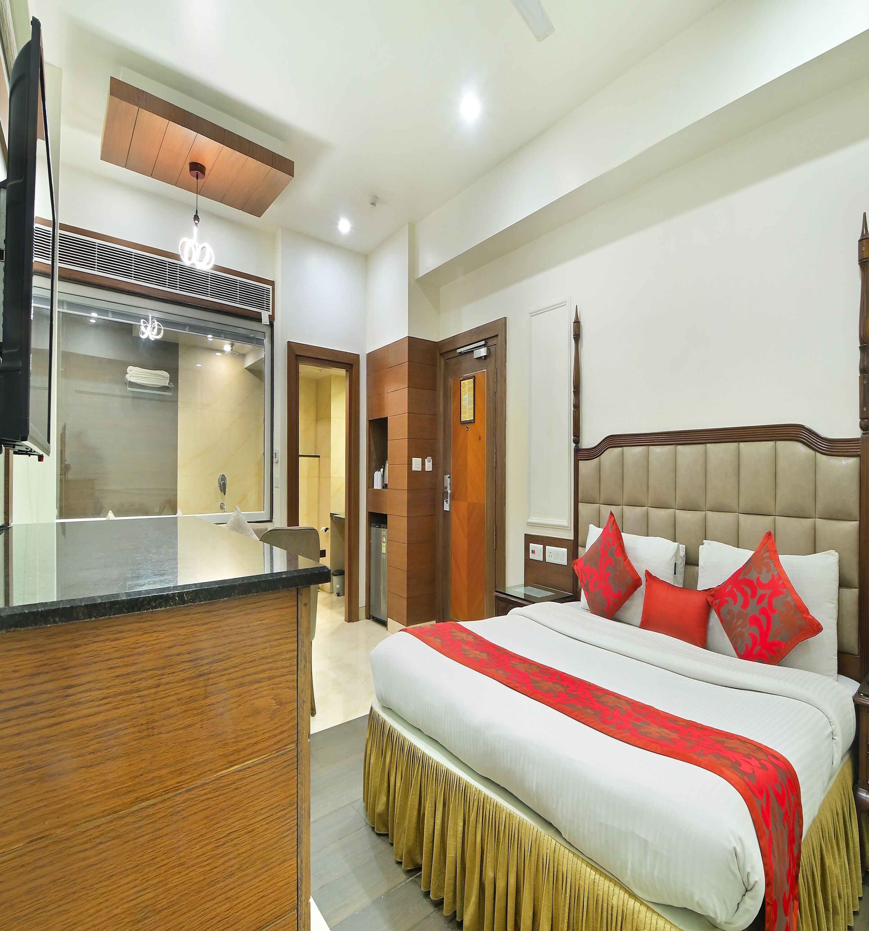 A hotel room with a bed, bedside table, and a mirror. There is a wooden desk and a chair in the corner.