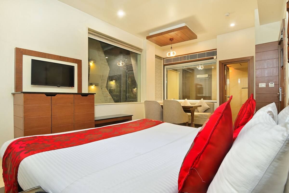 A hotel room with a large bed, a TV mounted on the wall, a desk with a chair, and a window with a view of the city.