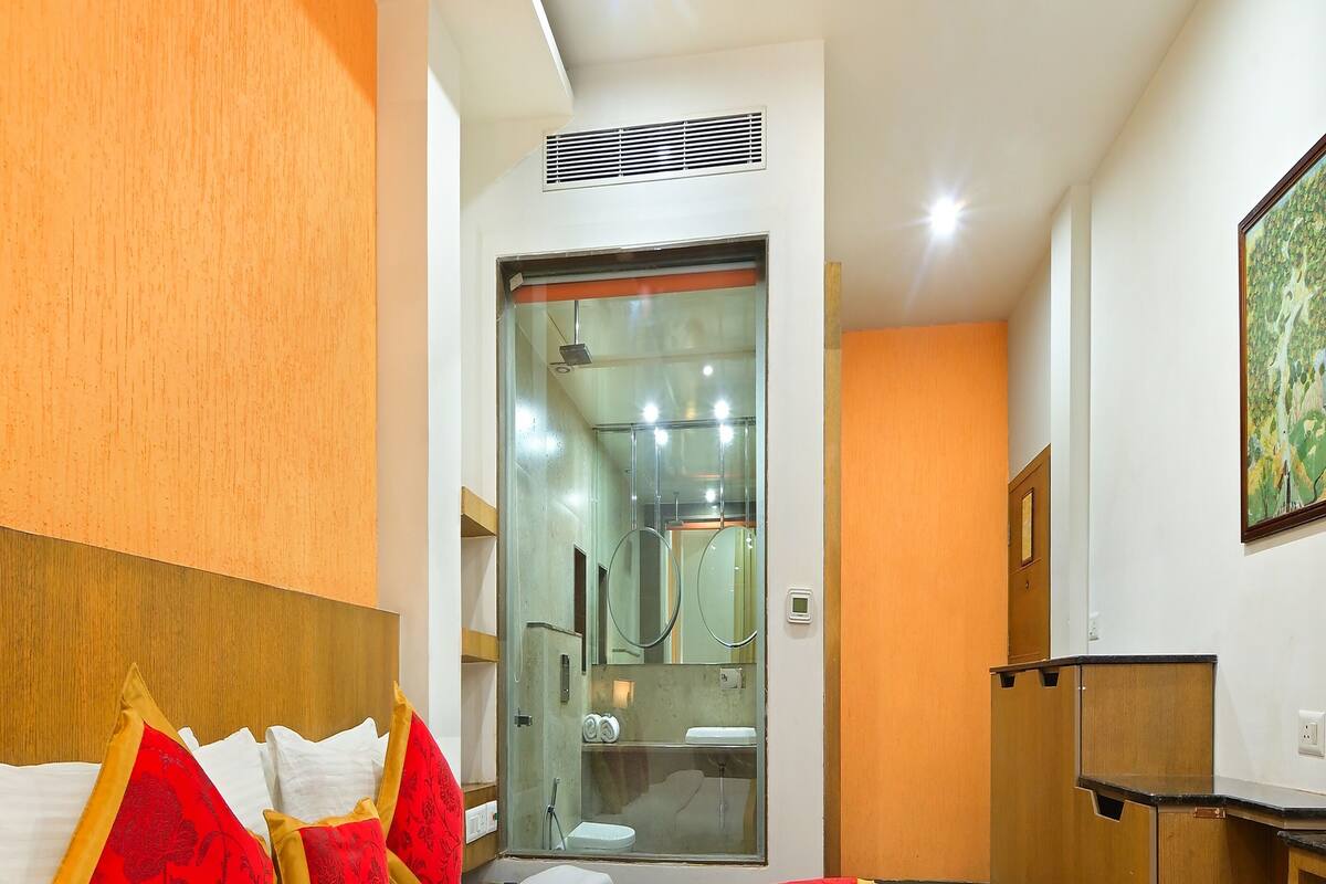 A hotel room with a bed, a wooden headboard, a desk, and a bathroom visible through a glass door.