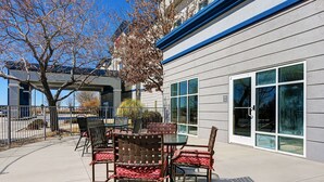 An outdoor patio with a table and chairs, a tree, and a building with large windows and a blue trim. - Hampton Inn Deming (Deming)