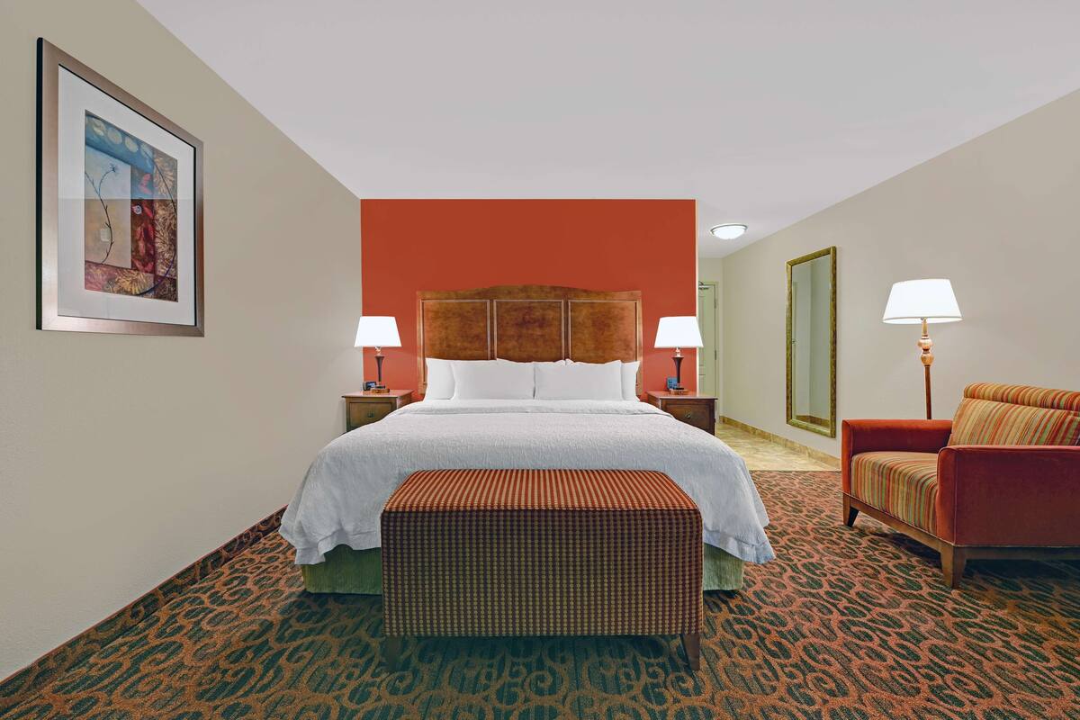 A hotel room with a large bed, two bedside tables, a chair, a bench, and a framed artwork on the wall.