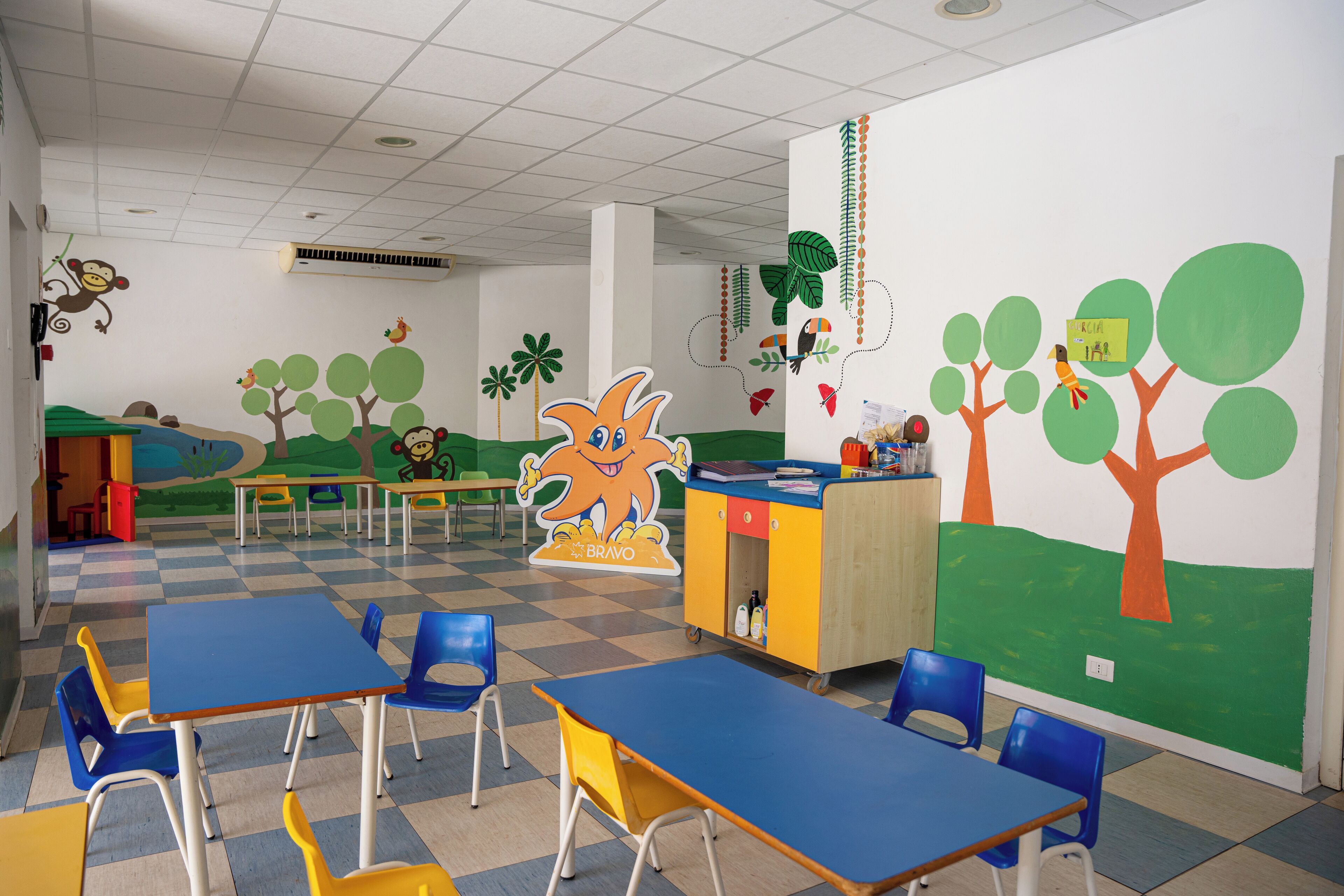 A classroom with blue and yellow tables, chairs, and a colorful mural on the wall.