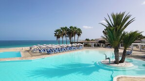 A pool area with lounge chairs and umbrellas, palm trees, and a view of the ocean. - VOI Praia de Chaves Resort - All inclusive (Boa Vista)