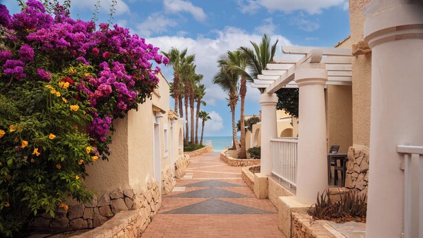A paved pathway flanked by white buildings with stone walls and palm trees, leading towards the sea. - VOI Praia de Chaves Resort - All inclusive (Boa Vista)