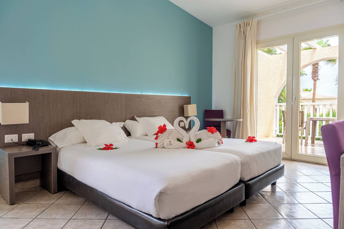A hotel room with two beds, a nightstand, a lamp, and a view of the outdoor balcony.