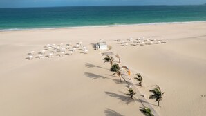 An aerial view of a beach with white umbrellas and lounge chairs arranged in rows, a pathway leading to a structure, and palm trees lining the path. - VOI Praia de Chaves Resort - All inclusive (Boa Vista)
