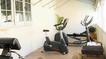 A covered patio area with exercise equipment, including an elliptical and a stationary bike, a bench, and a small table with towels.