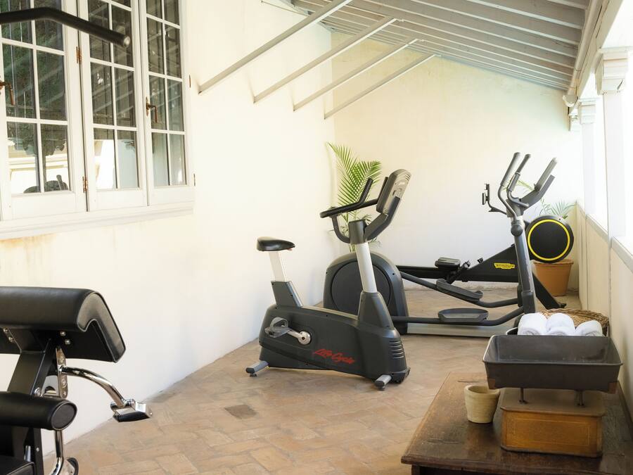A covered patio area with exercise equipment, including an elliptical and a stationary bike, a bench, and a small table with towels.