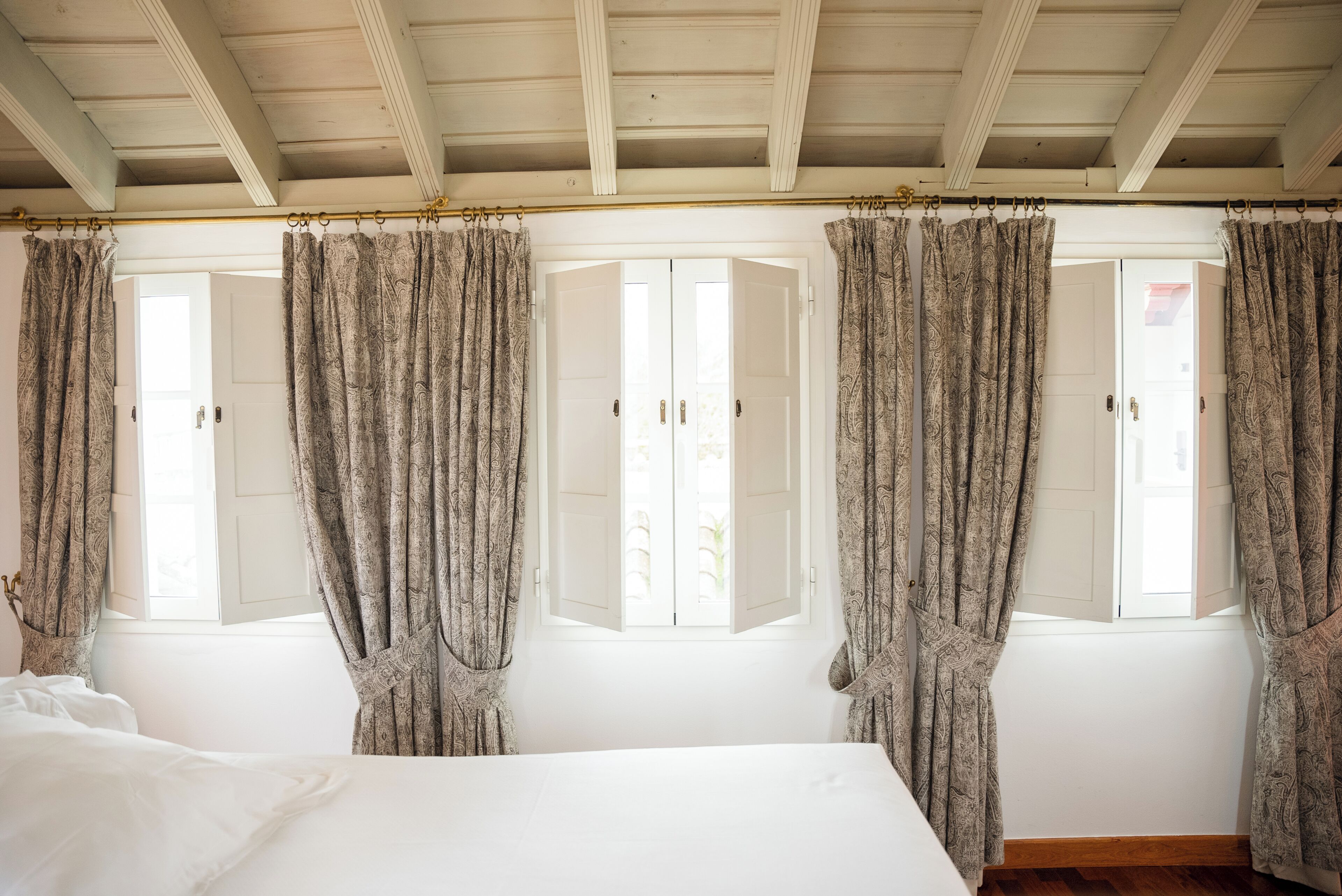 Une chambre à coucher avec un lit, deux fenêtres avec volets et des rideaux à motifs.