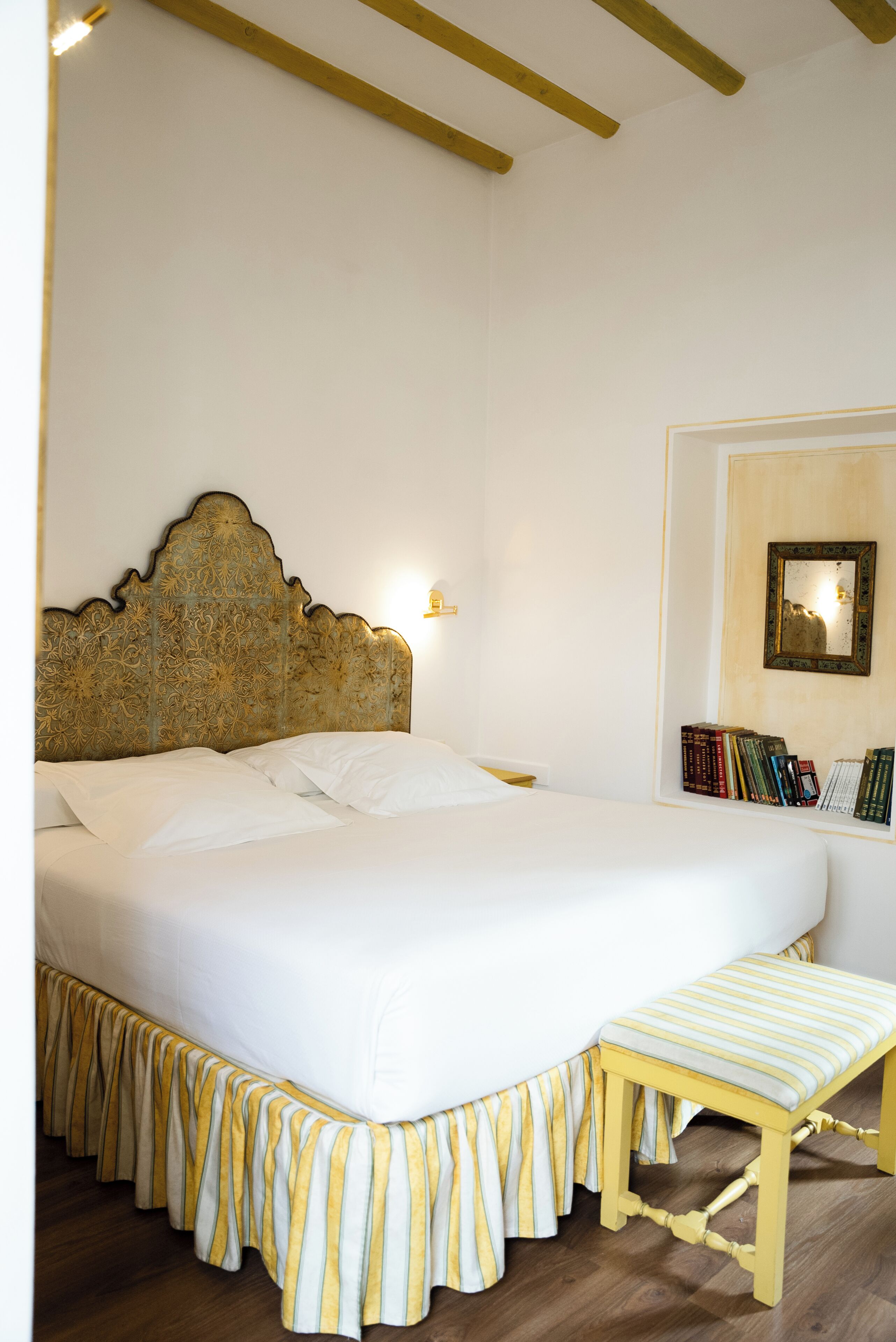 A bedroom with a large bed, a bedside table, a wall-mounted lamp, and a bookshelf.