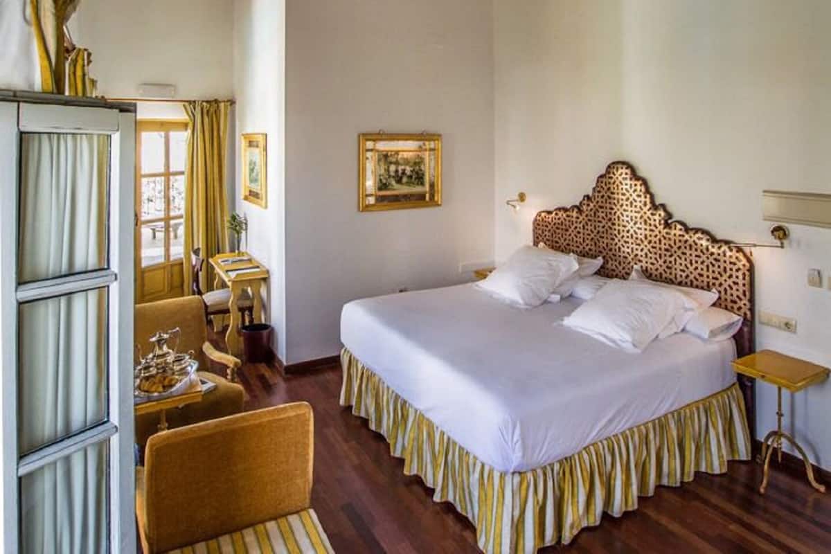 A bedroom with a large bed, a wooden chair, a desk, and a ceiling with wooden beams and windows.