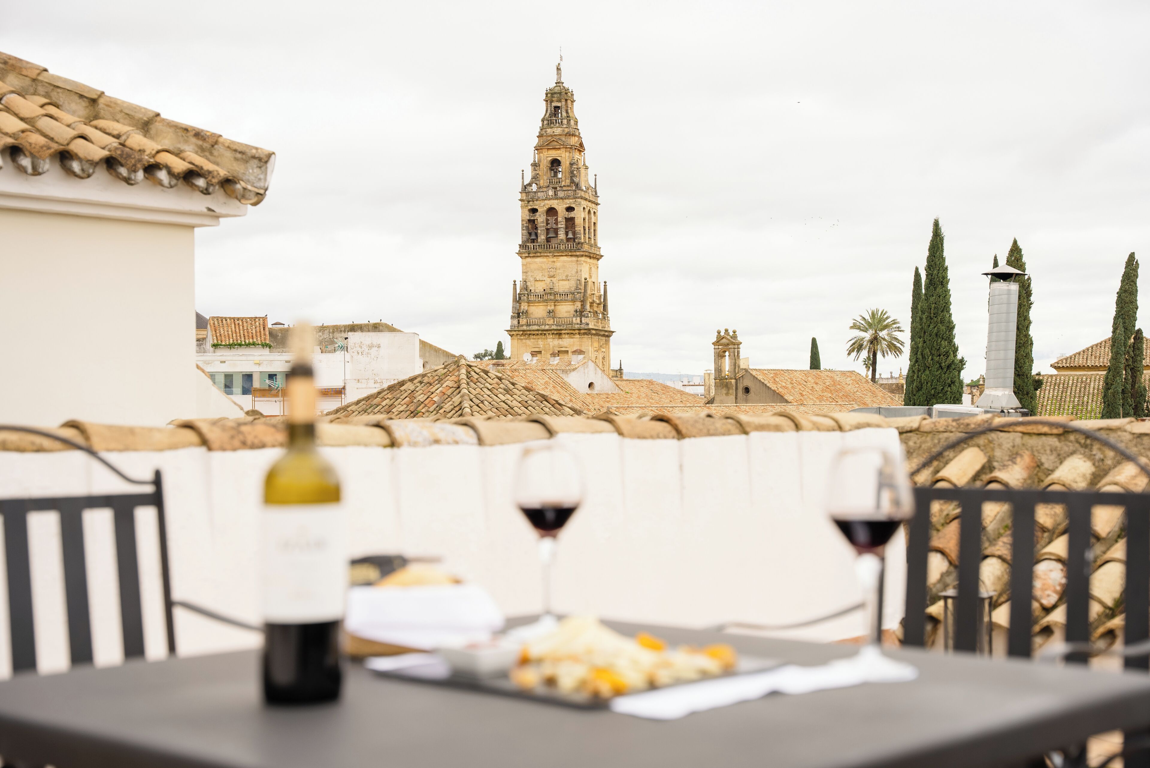 Una terraza con una mesa, dos copas de vino y una botella. Al fondo, una torre histórica y una palmera.