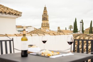 A terrace with a table, two wine glasses, and a bottle of wine. In the background, there is a historic tower and a palm tree.