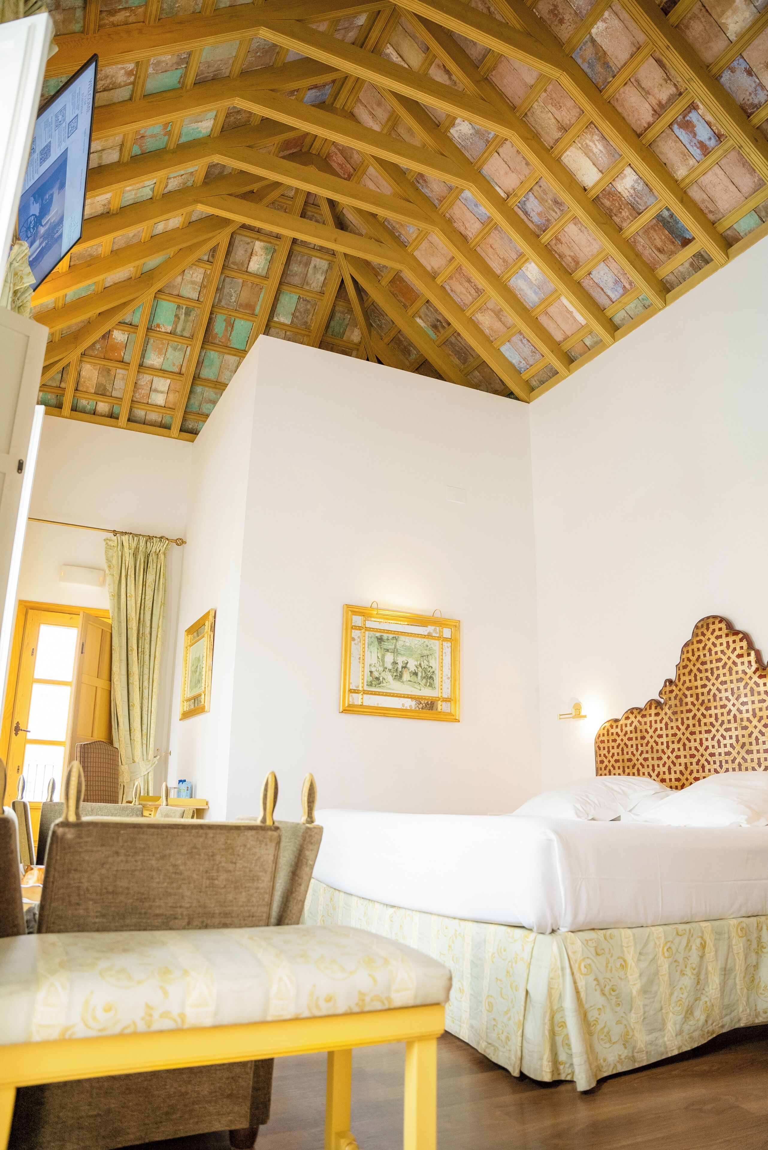 A hotel room with a high wooden ceiling, a bed, a bedside table, a chair, and a wall-mounted picture.