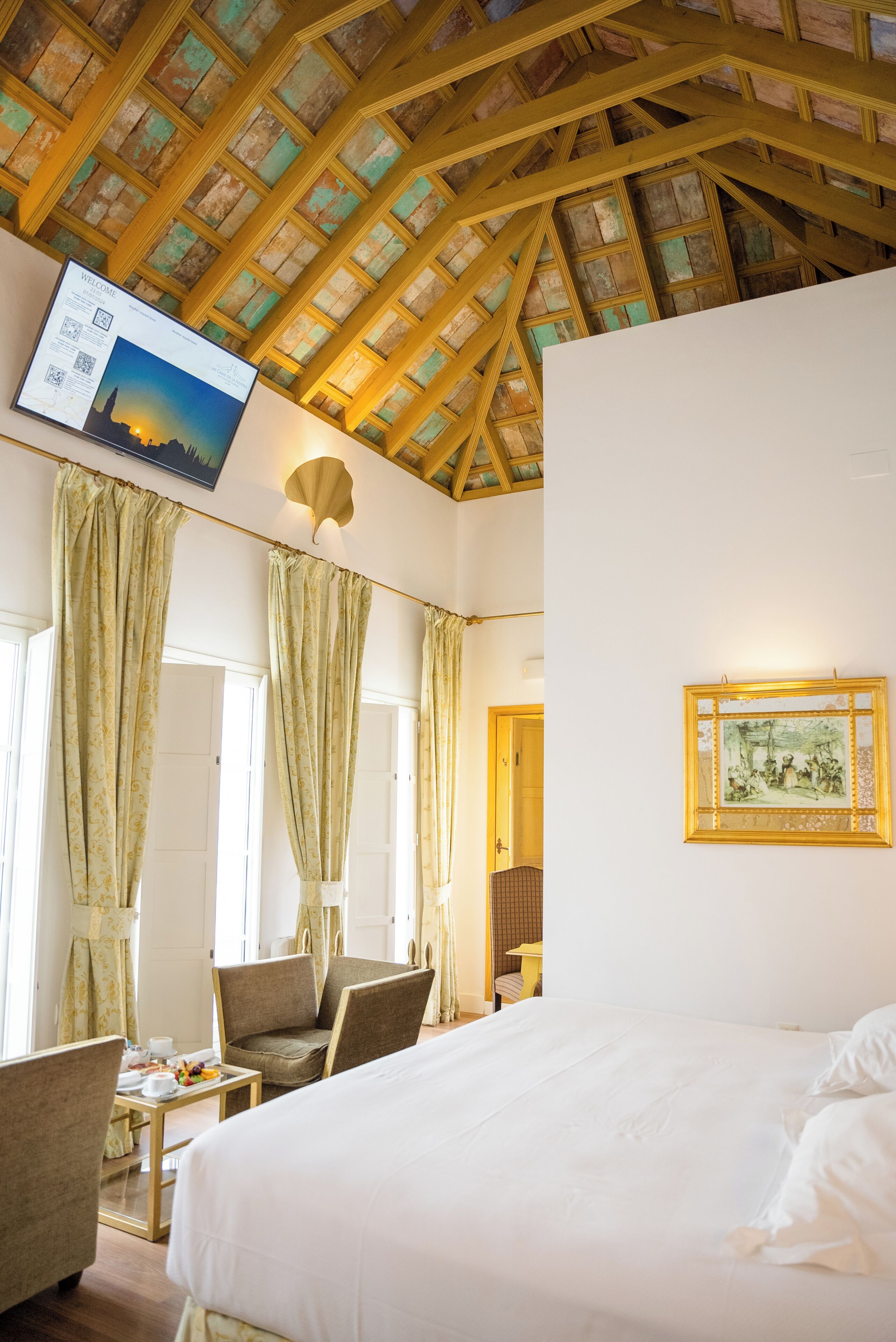 A hotel room with a large bed, two chairs, a small table with breakfast items, a framed picture on the wall, and a ceiling with wooden beams.