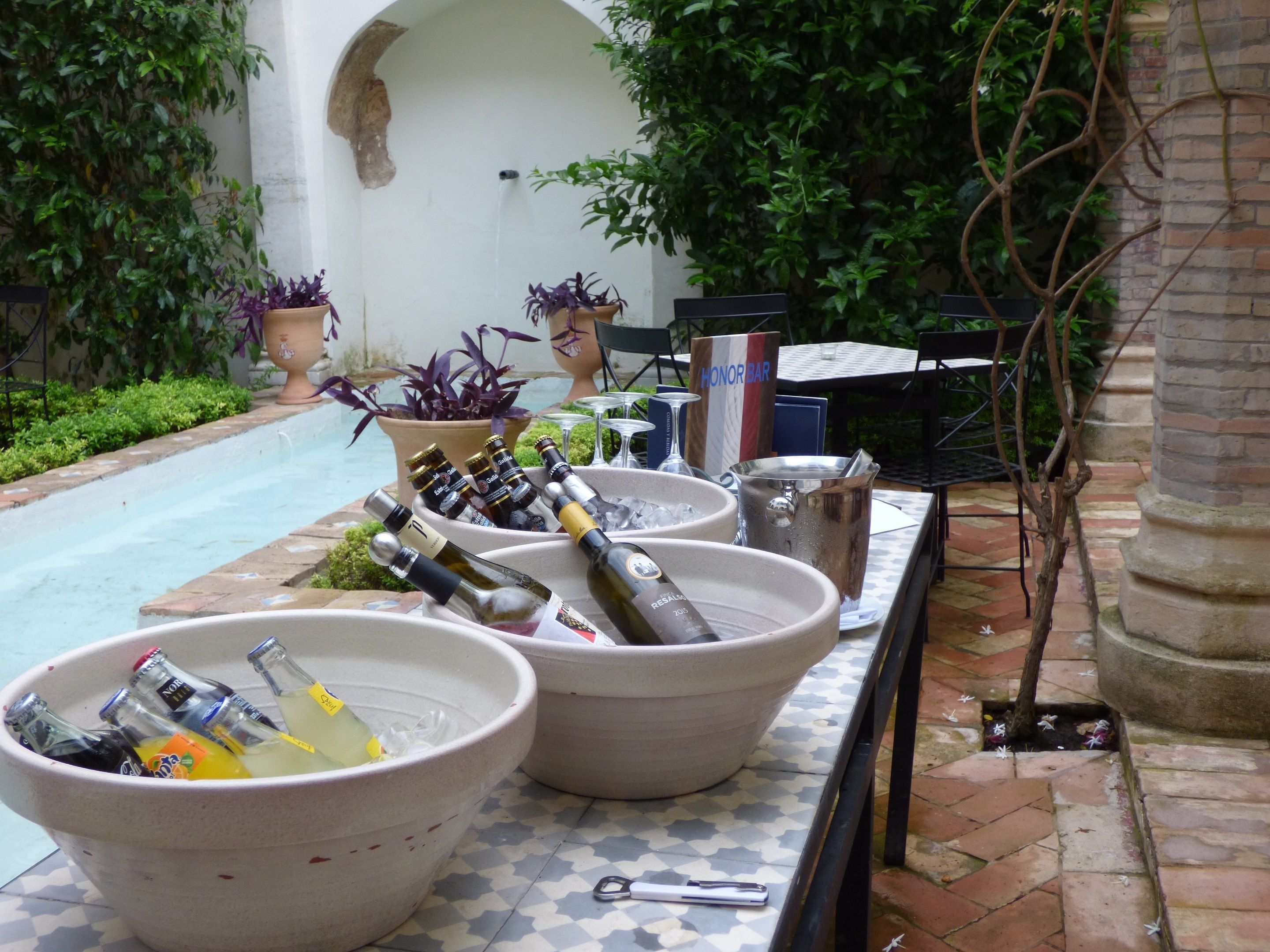 Un espace bar aménagé au bord de la piscine, avec de grands récipients remplis de glace et des bouteilles de boissons, entouré de plantes en pot et de mobilier d’extérieur.