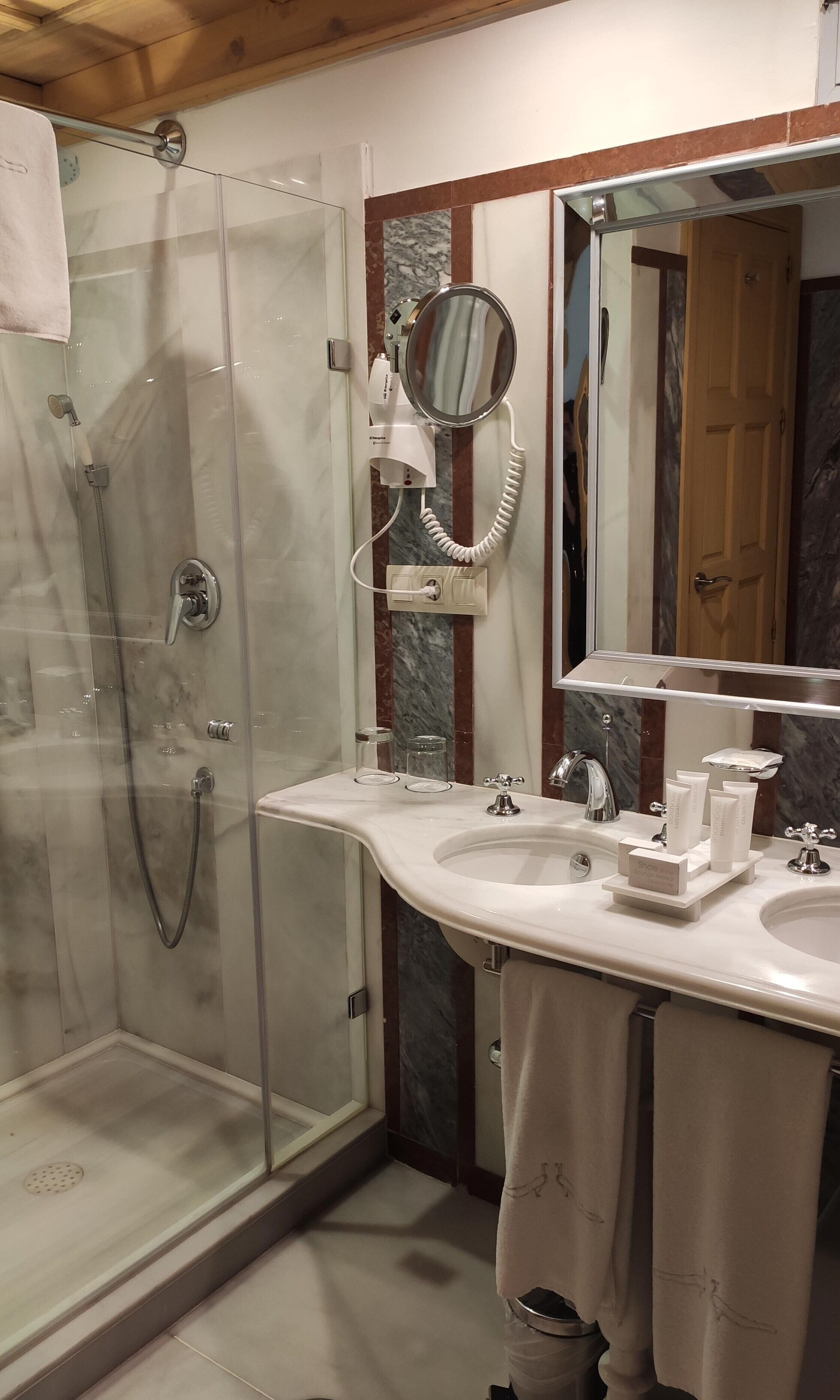 Un baño moderno con ducha de vidrio, tocador con dos lavamanos, un espejo grande y un secador de pelo en la pared.