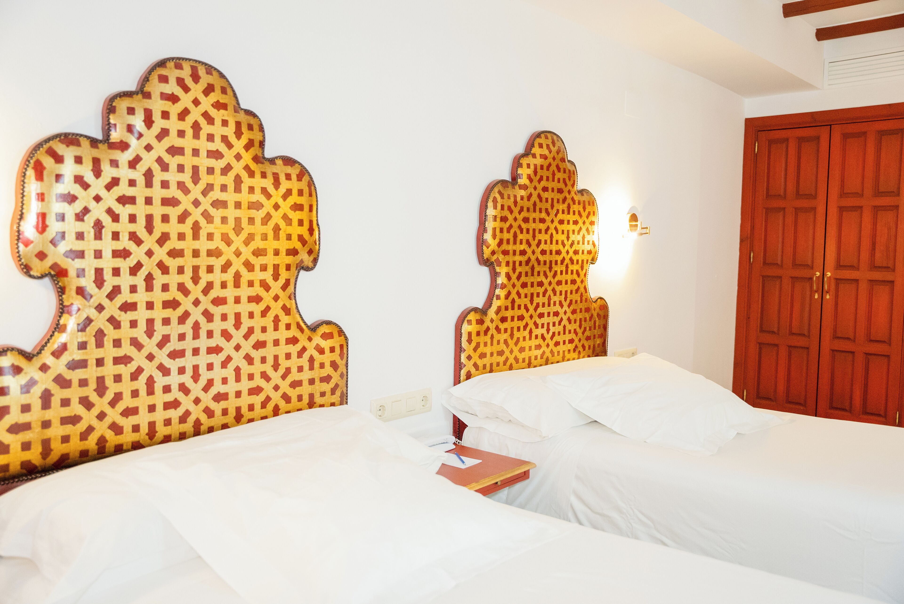 Une chambre d’hôtel avec deux lits, chacun doté d’une tête de lit à motifs, d’une table de chevet avec un livre et d’une armoire en bois.