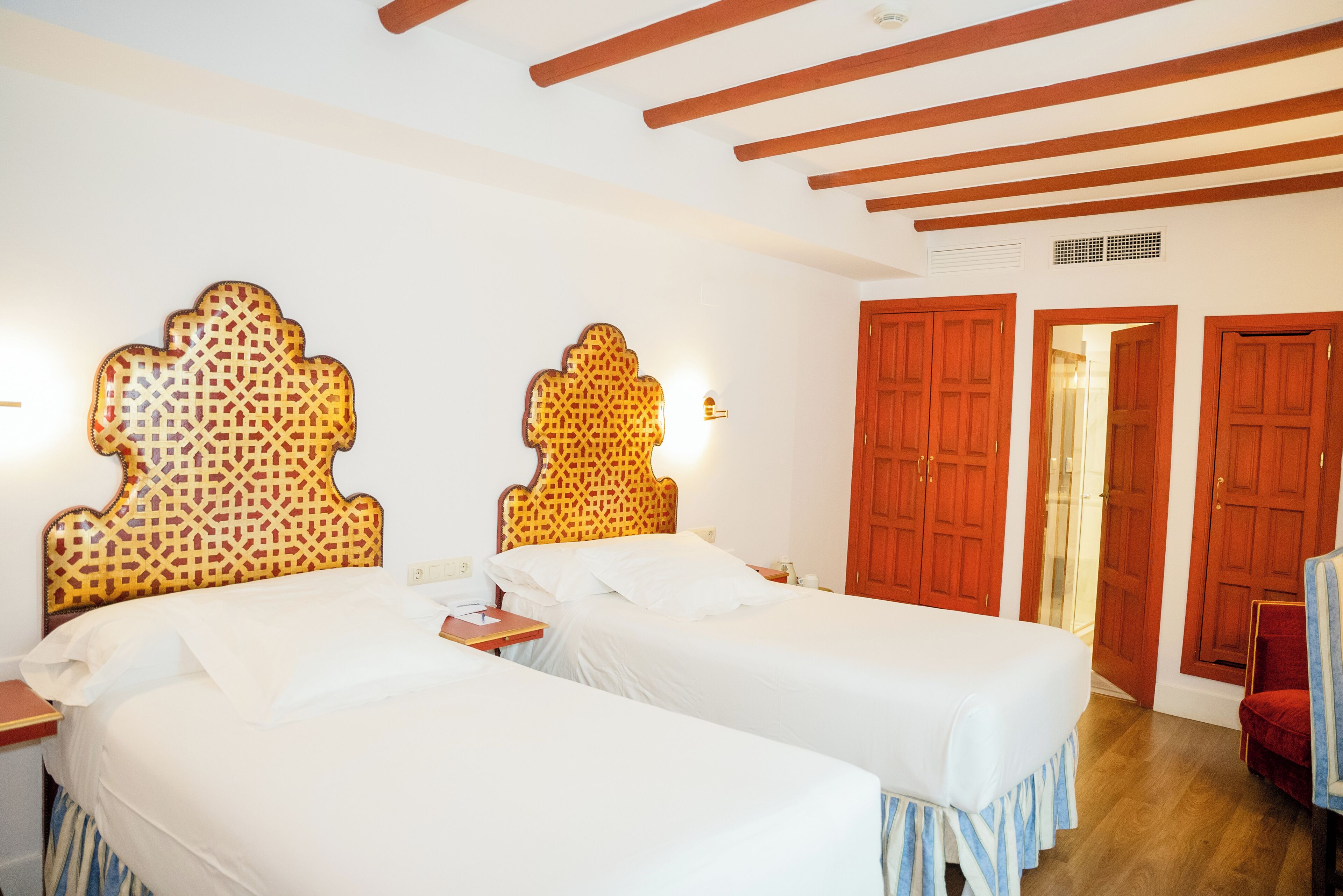 Habitación de hotel con dos camas individuales, cabeceros de madera, una mesita de noche y una silla roja.