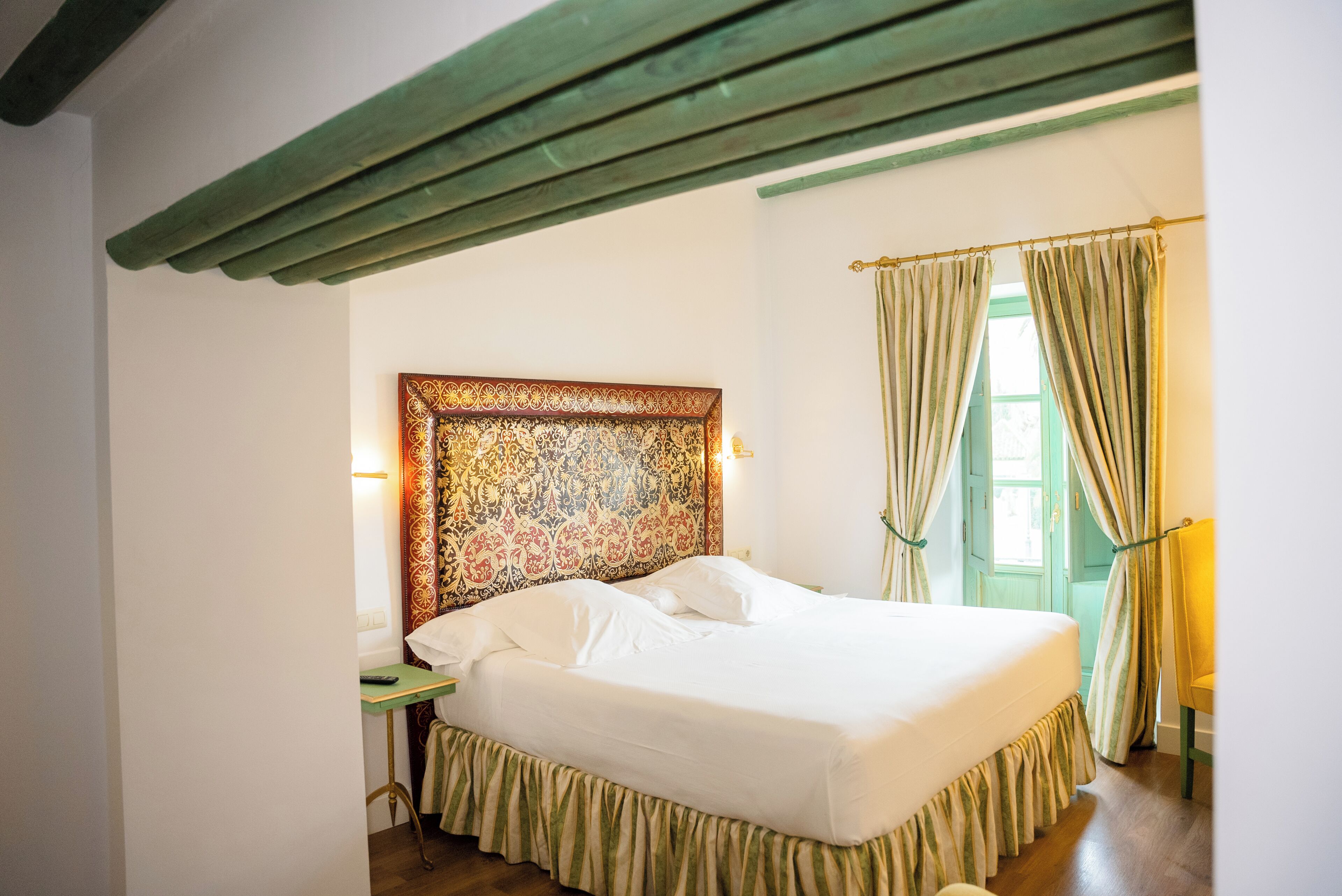 A room with a bed, a nightstand, a window with curtains, and a wall with a decorative headboard.