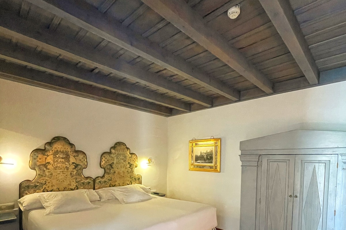 A bedroom with a wooden ceiling, a bed with a striped skirt, a bench, a nightstand, a wall-mounted lamp, and a wardrobe.